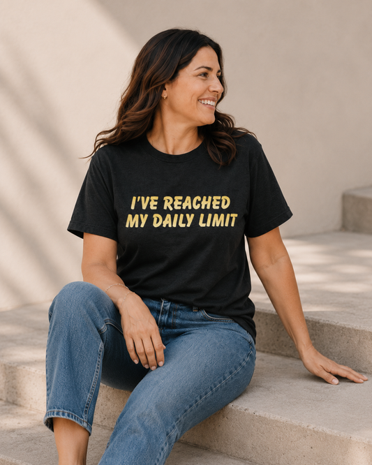 Woman wearing black 'I'VE REACHED MY DAILY LIMIT' slogan t-shirt, alternate pose against a tan wall