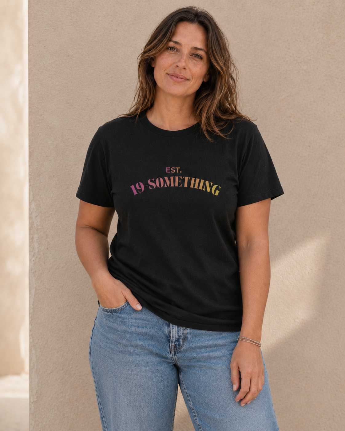 Woman wearing black t-shirt with 'EST. 19-SOMETHING' slogan in pink-to-yellow gradient text, standing against a tan wall