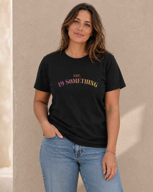 Woman wearing black t-shirt with 'EST. 19-SOMETHING' slogan in pink-to-yellow gradient text, standing against a tan wall