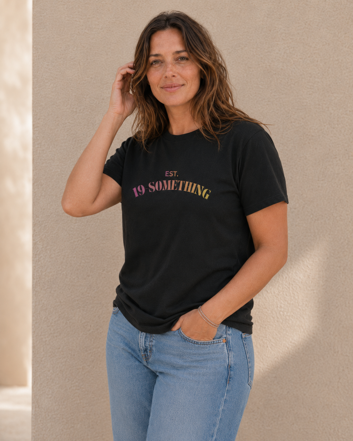 Woman wearing black 'EST. 19-SOMETHING' slogan t-shirt, alternate pose against a tan wall