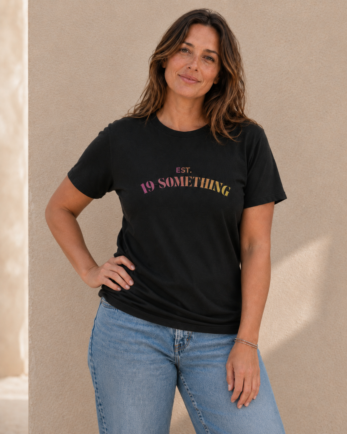 Close-up of woman wearing black 'EST. 19-SOMETHING' slogan t-shirt against a tan wall