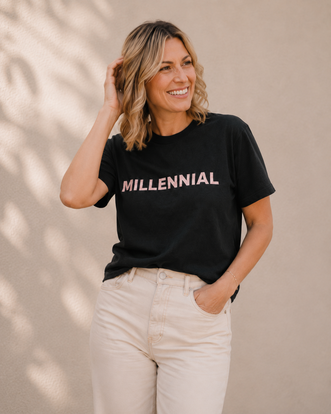 Woman wearing black 'MILLENNIAL' slogan t-shirt, alternate pose against a tan wall