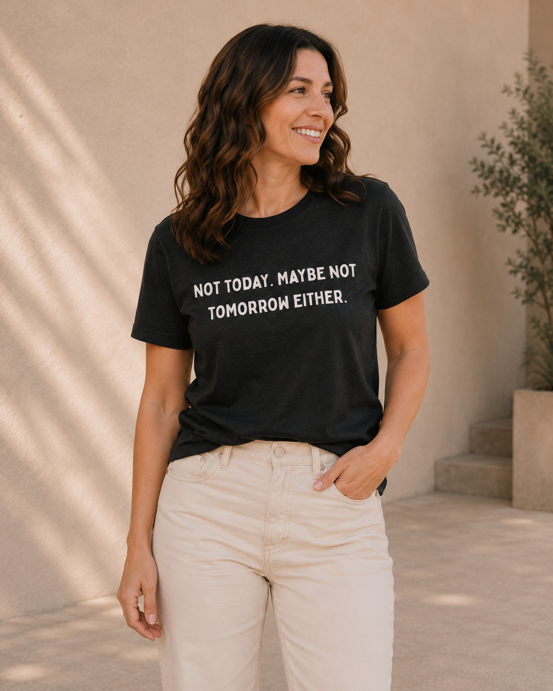 Woman wearing black 'NOT TODAY. MAYBE NOT TOMORROW EITHER' slogan t-shirt, alternate pose against a tan wall