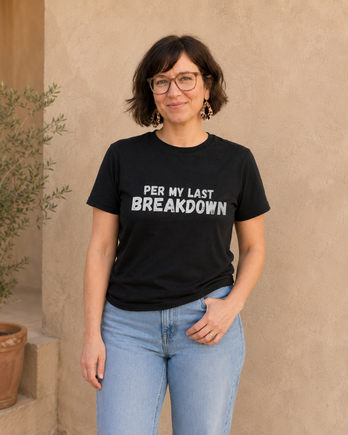 Woman wearing black 'PER MY LAST BREAKDOWN' slogan t-shirt, alternate pose against a tan wall