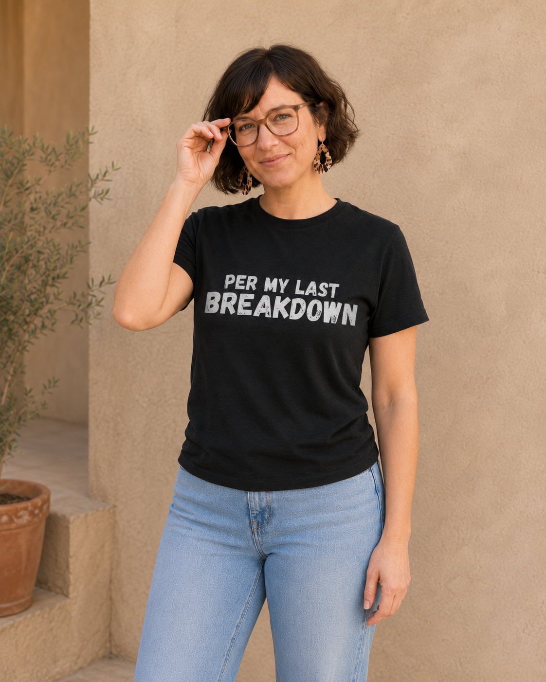 Woman wearing black t-shirt with white 'PER MY LAST BREAKDOWN' slogan text, standing against a tan wall