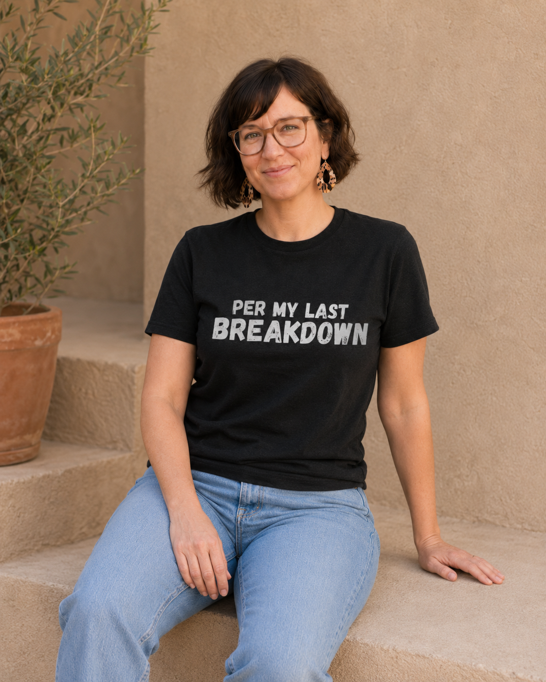 Close-up of woman wearing black 'PER MY LAST BREAKDOWN' slogan t-shirt against a tan wall