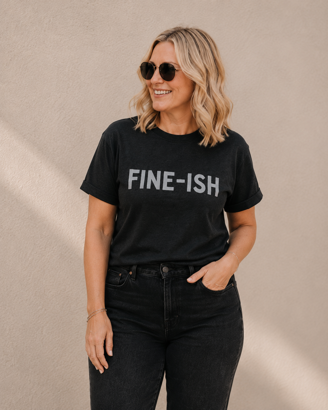 Woman wearing black 'FINE-ISH' slogan t-shirt, alternate pose against a tan wall