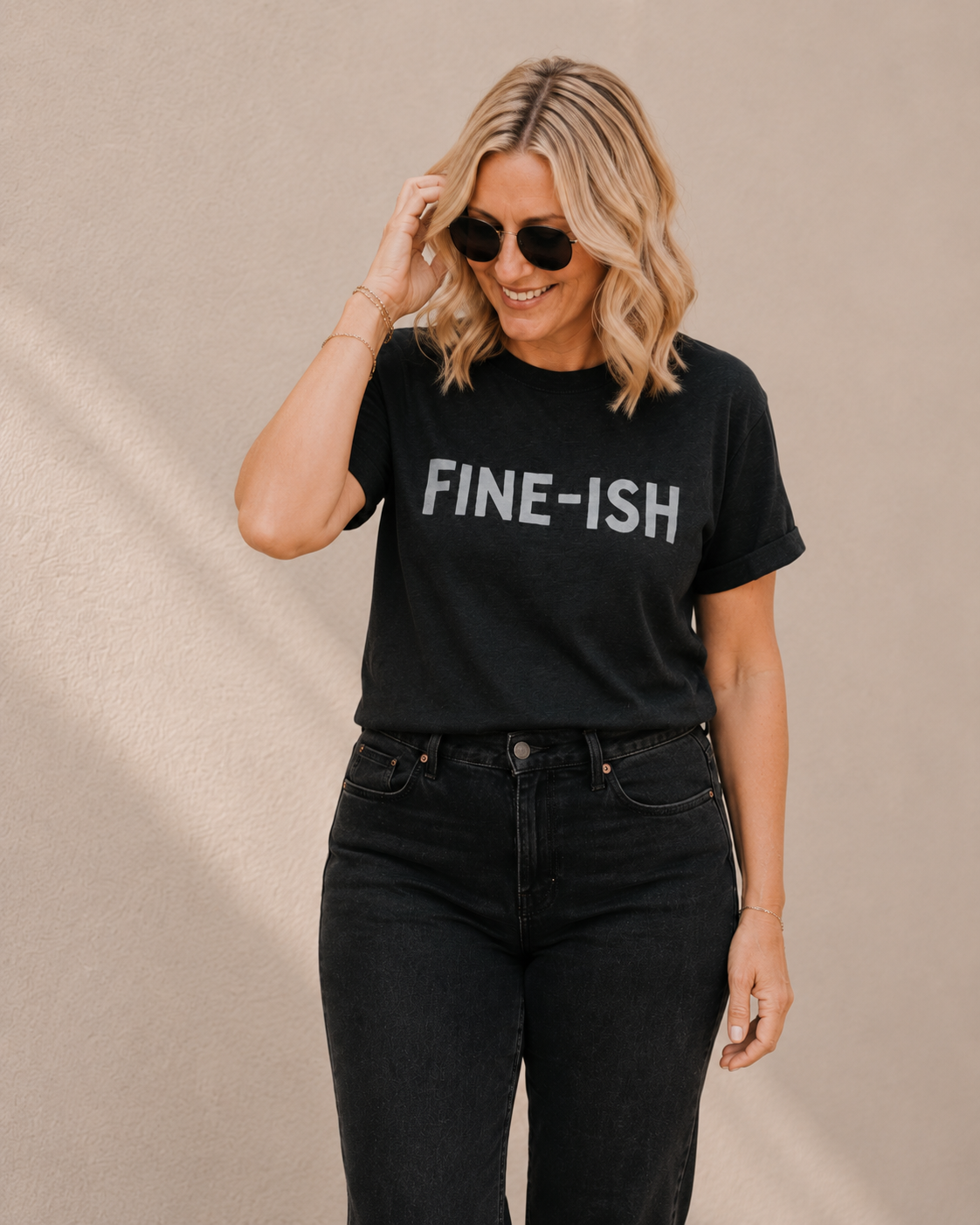 Woman wearing black t-shirt with 'FINE-ISH' slogan text, standing against a tan wall