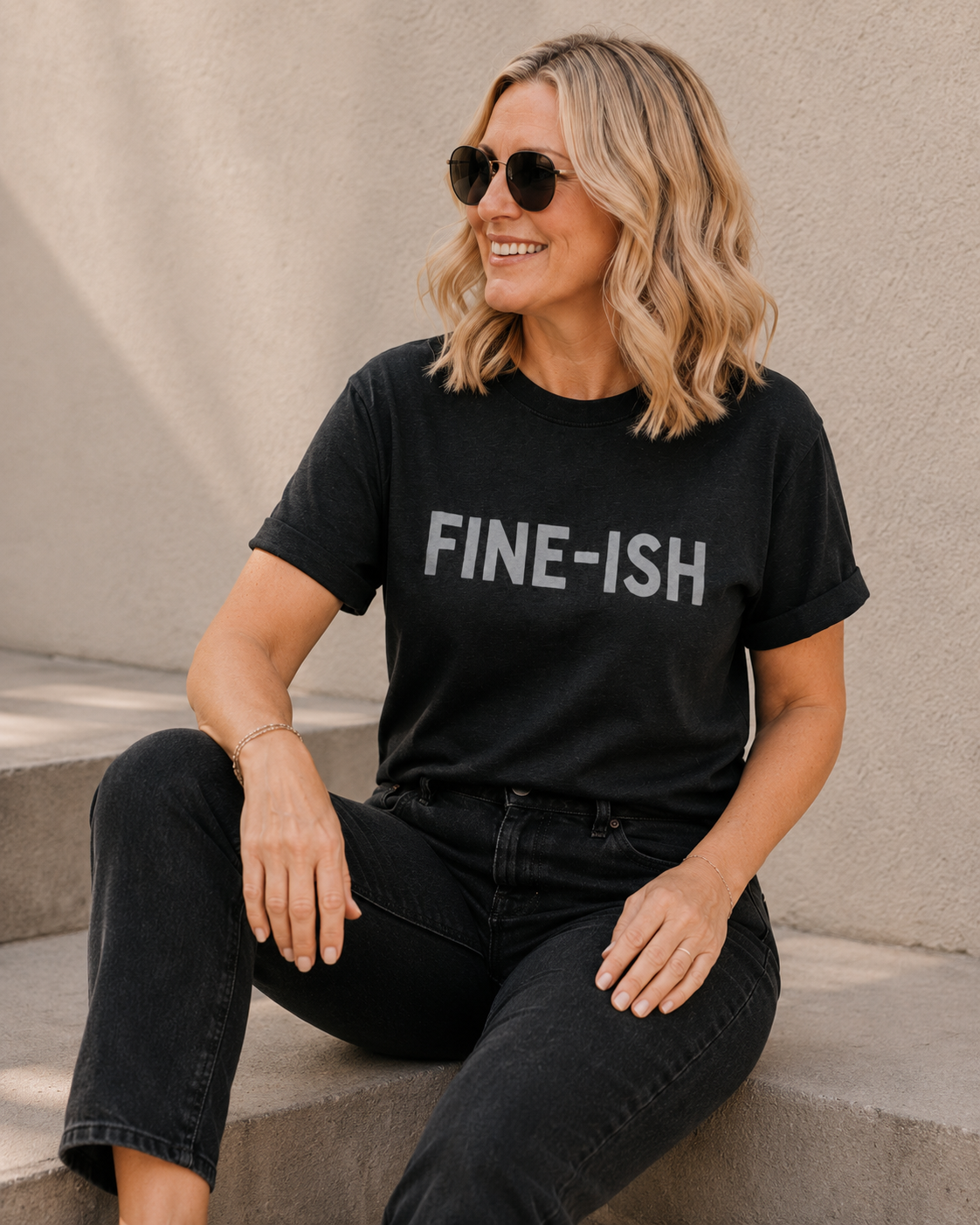 Close-up of woman wearing black 'FINE-ISH' slogan t-shirt against a tan wall