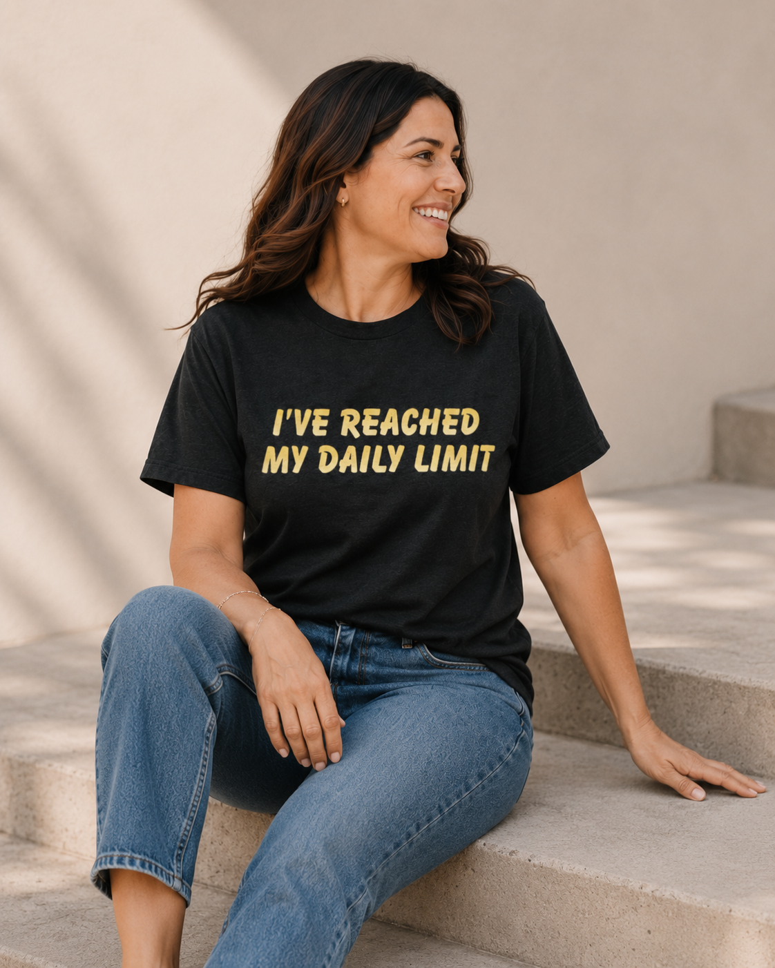 Woman wearing black 'I'VE REACHED MY DAILY LIMIT' slogan t-shirt, alternate pose against a tan wall