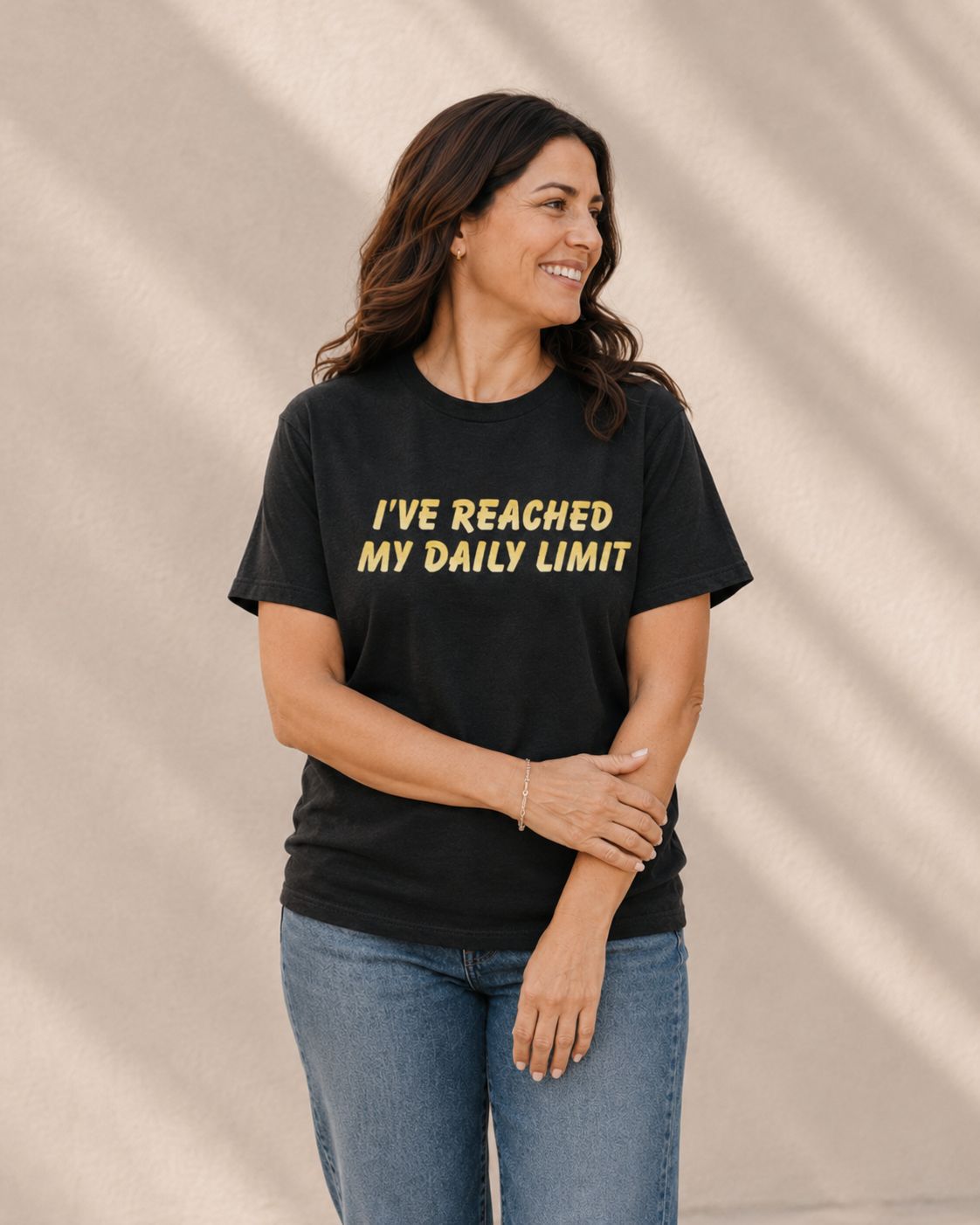 Close-up of woman wearing black 'I'VE REACHED MY DAILY LIMIT' slogan t-shirt against a tan wall