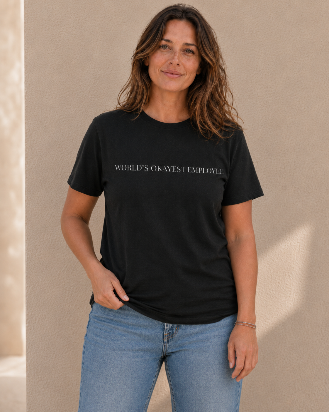 Woman wearing black t-shirt with white 'WORLD'S OKAYEST EMPLOYEE' slogan text, standing against a tan wall