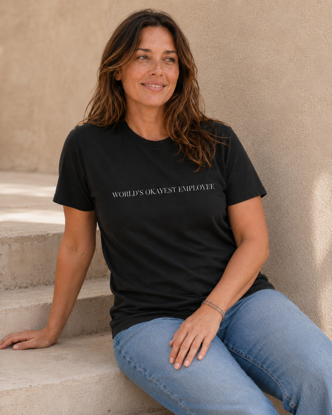 Woman wearing black 'WORLD'S OKAYEST EMPLOYEE' slogan t-shirt, alternate pose against a tan wall
