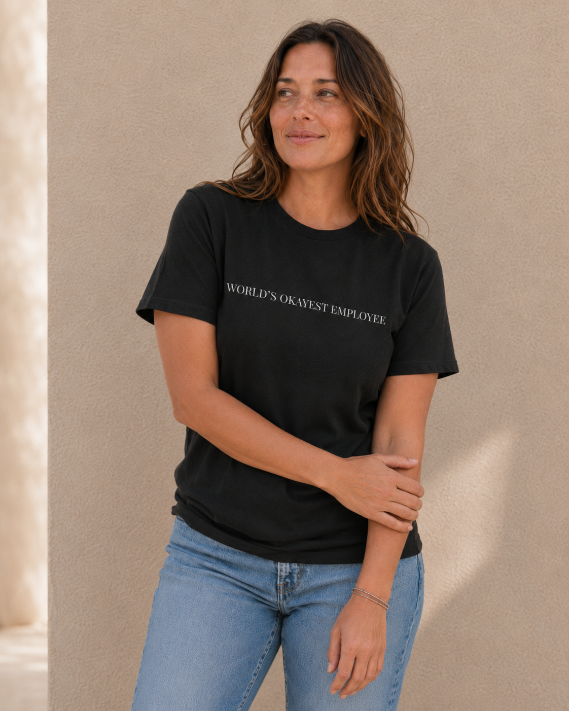 Close-up of woman wearing black 'WORLD'S OKAYEST EMPLOYEE' slogan t-shirt against a tan wall