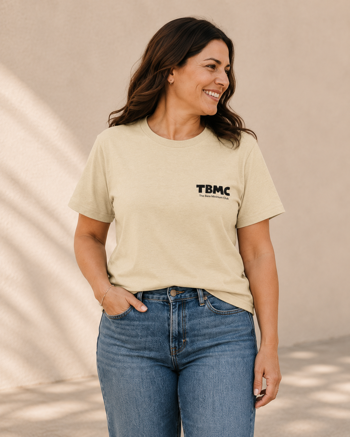 Close-up of woman wearing butter yellow TBMC logo t-shirt against a tan wall