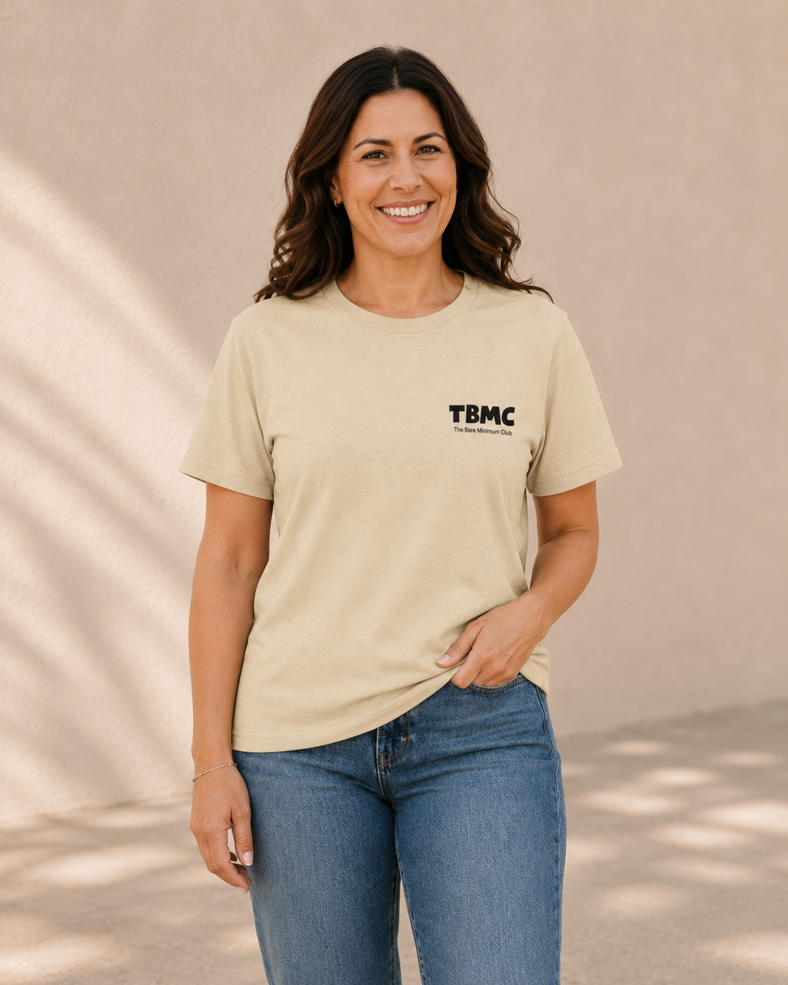 Woman wearing butter yellow t-shirt with small TBMC logo, standing against a tan wall