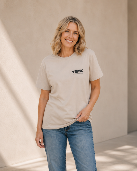 Woman wearing vintage white t-shirt with small TBMC logo, standing against a tan wall