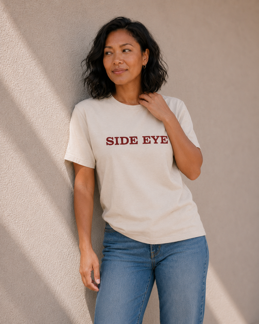 Woman wearing The Bare Minimum Club's vintage white 'Side Eye' graphic t-shirt, looking to the side against a sunlit wall