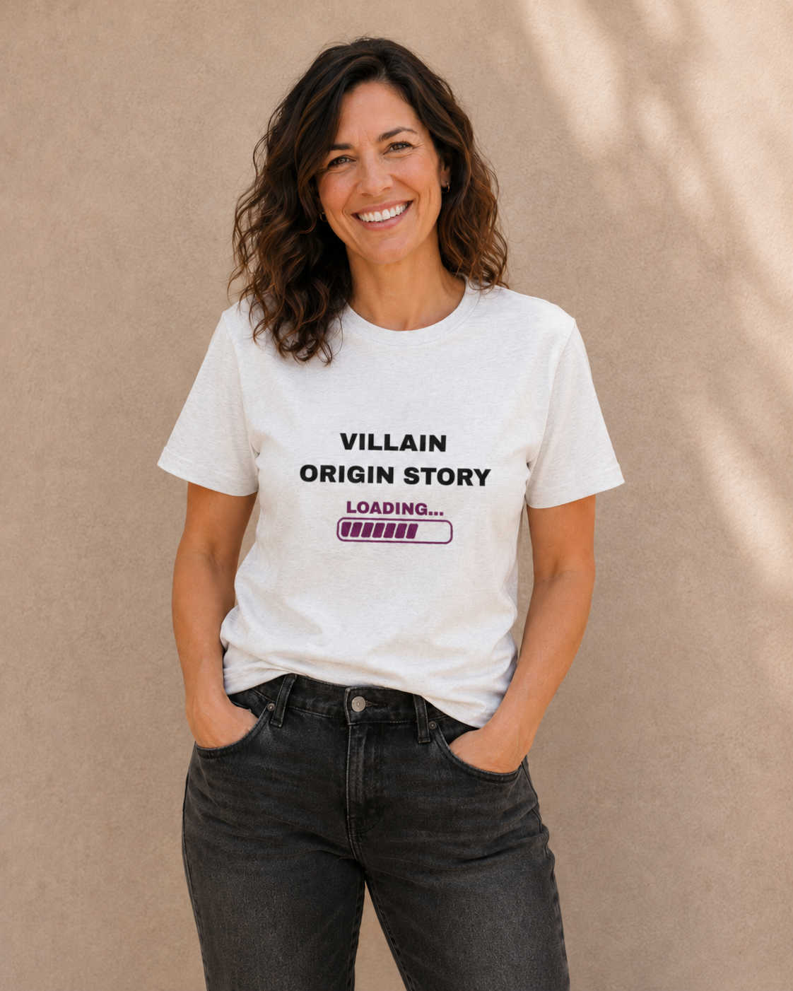 Woman wearing The Bare Minimum Club's white 'Villain Origin Story' graphic t-shirt, hands in pockets, standing against a sunlit wall