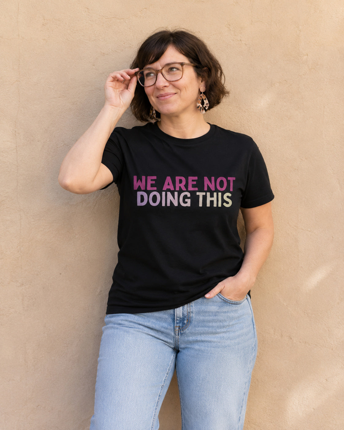 Woman wearing black t-shirt with 'WE ARE NOT DOING THIS' slogan in pink-to-lavender gradient text, standing against a tan wall