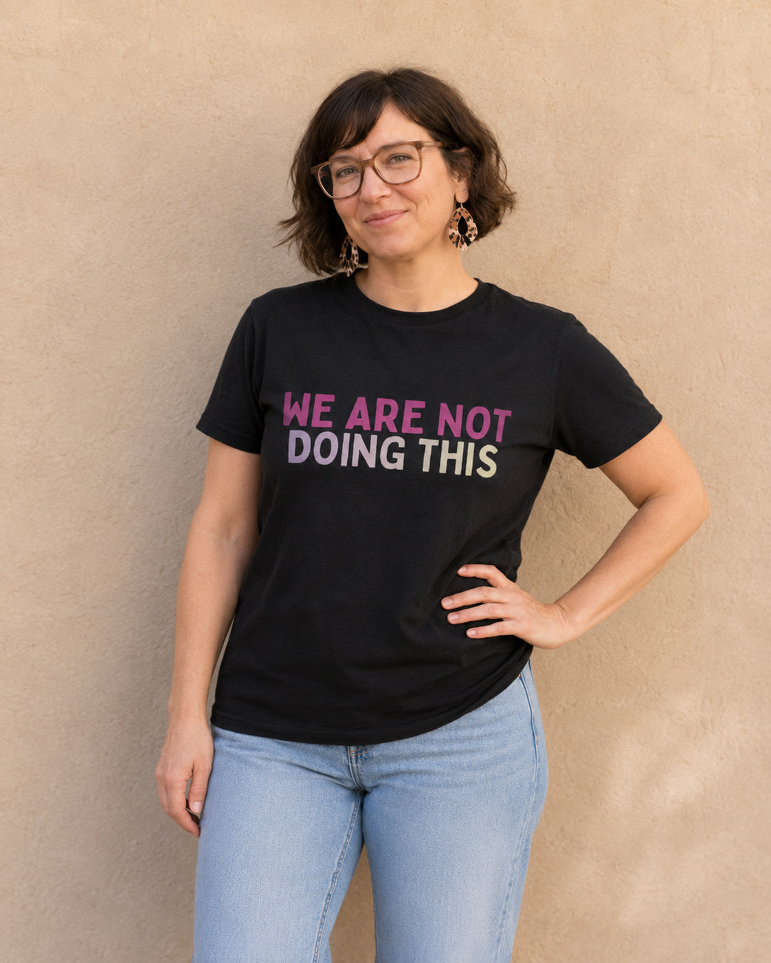Woman wearing black 'WE ARE NOT DOING THIS' slogan t-shirt, alternate pose against a tan wall