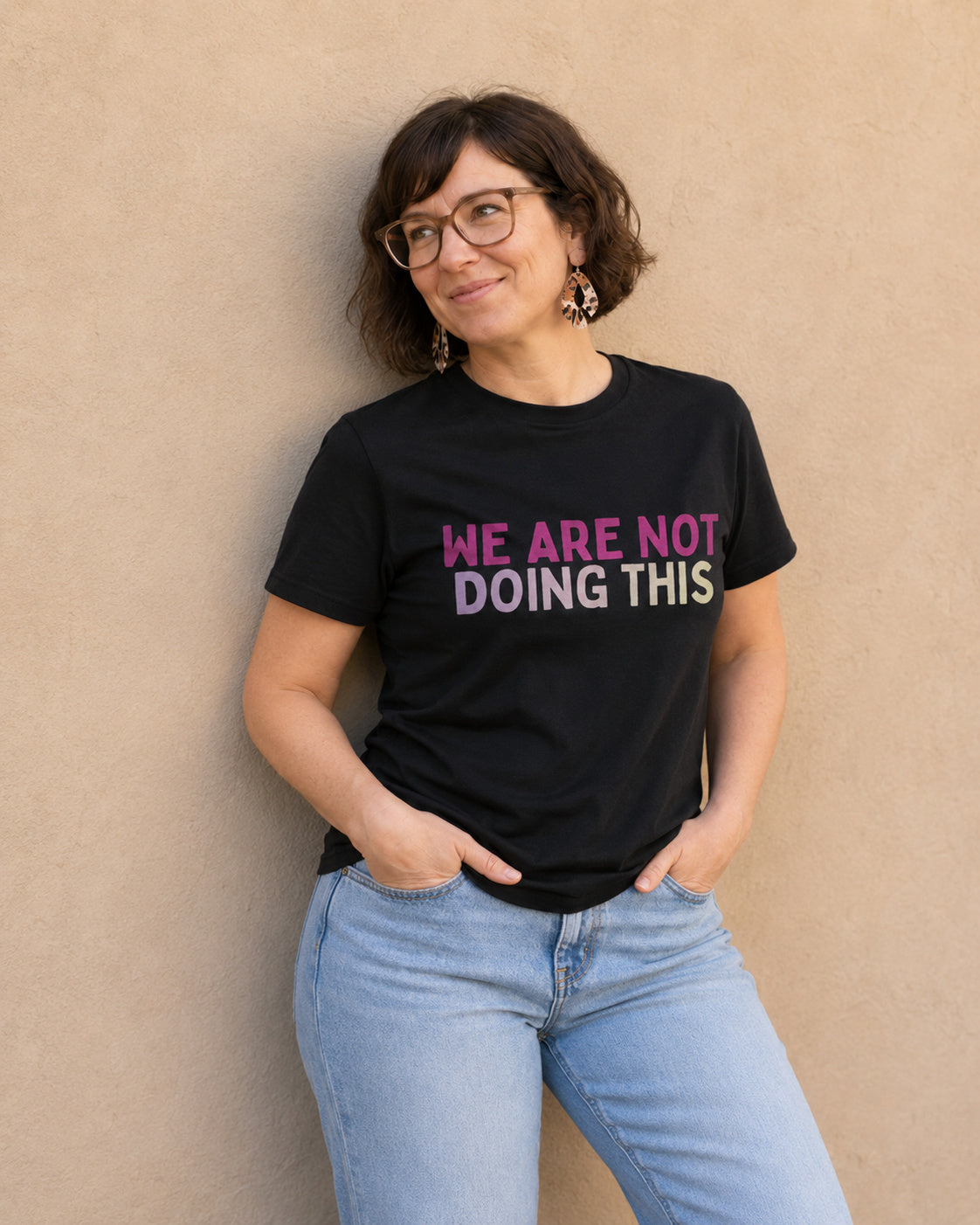 Close-up of woman wearing black 'WE ARE NOT DOING THIS' slogan t-shirt against a tan wall